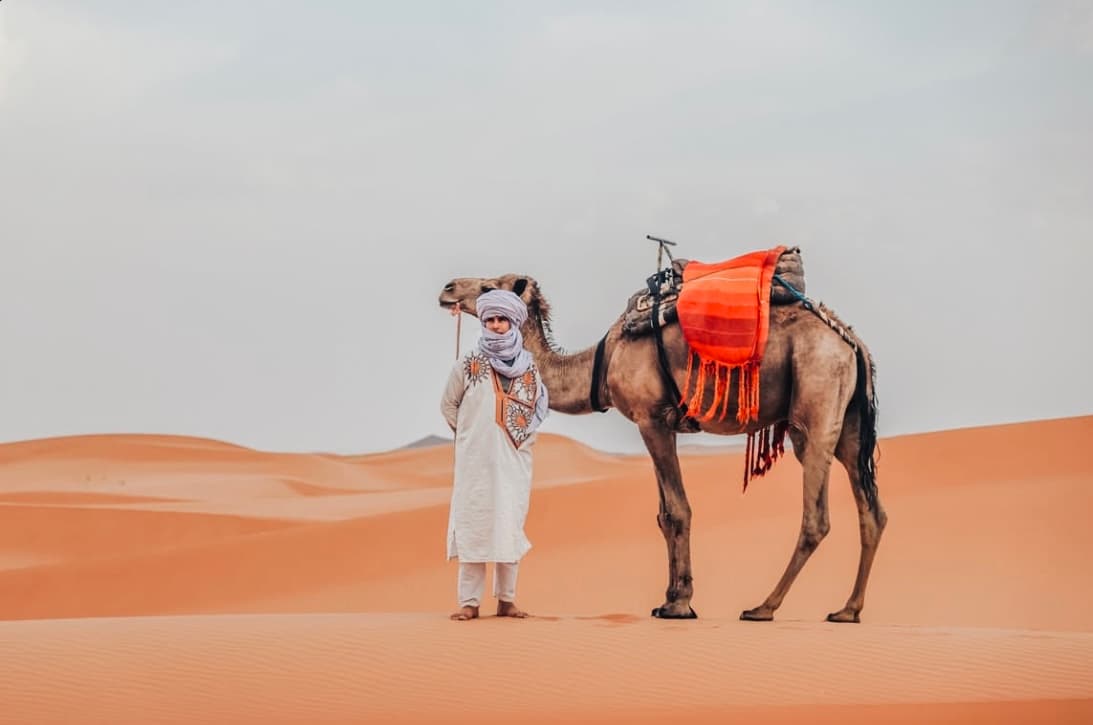 Younes in traditional desert clothing with a camel in the Sahara dunes